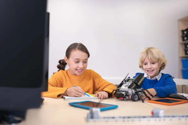 Μαθητές καθισμένοι στο γραφείο, κρατώντας σημειώσεις, παίζοντας με το κιτ του κτιρίου, χαμογελώντας — Φωτογραφία Αρχείου