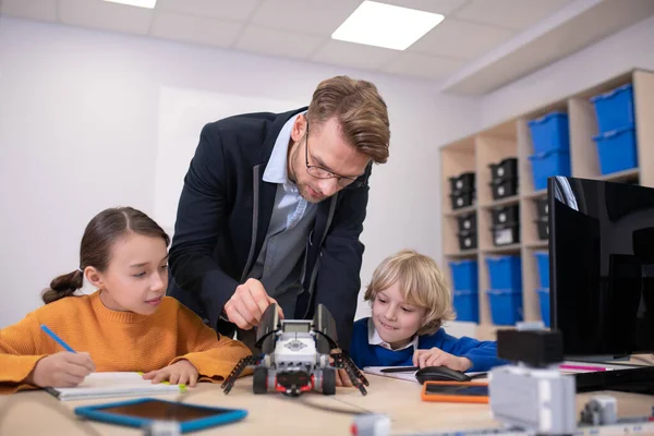 Pupils sitting at desk, playing with buildable car, male teacher explaining — Stock Photo, Image