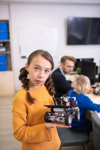 Girl holding buildable car, teacher and boy sitting in background