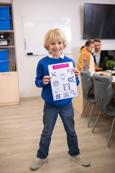 Ξανθό αγόρι στέκεται και κρατώντας χαρτί με σχέδια της φυσικής — Φωτογραφία Αρχείου