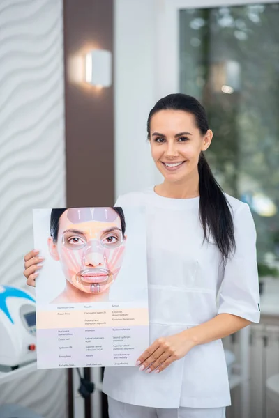 Cosmetólogo sonriente sosteniendo papel con información sobre la cara — Foto de Stock