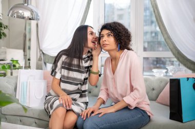 Two dark-haired women sitting on the sofa andsharing gossips