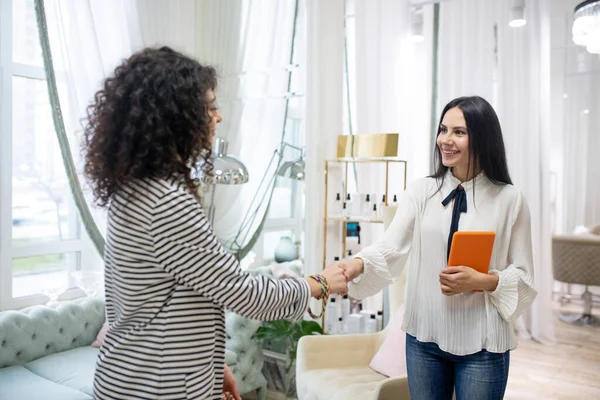 Curly-haired beautiful customer in a striped jacket shaking hands with beautician — 스톡 사진