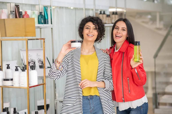 Dos chicas guapas de cabello oscuro mostrando productos para el cuidado del cuerpo y sonriendo — Foto de Stock