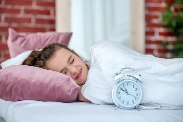 Gadis tidur di tempat tidur putih, sebelah jam alarm putih . — Stok Foto