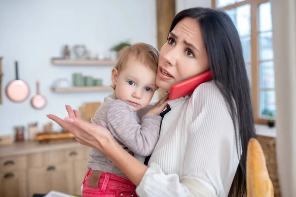 Работающая мама держит ребенка, разговаривает по телефону, чувствует себя расстроенной — стоковое фото