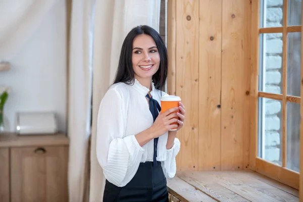 Jonge vrouw die voor het raam staat, koffiebeker vasthoudt, glimlacht — Stockfoto