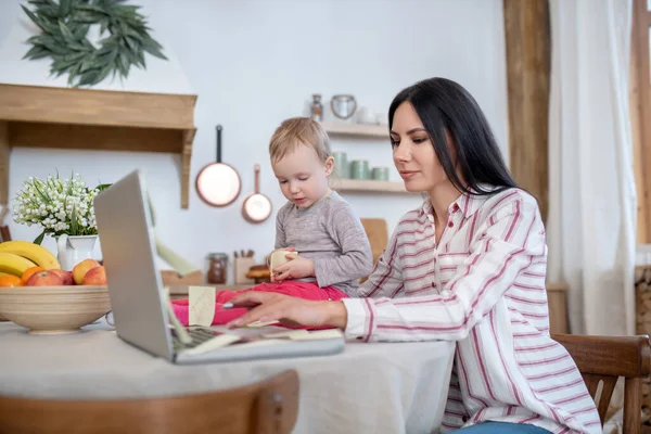 Joven mamá escribiendo en el ordenador portátil, hija sentada a su lado — Foto de Stock