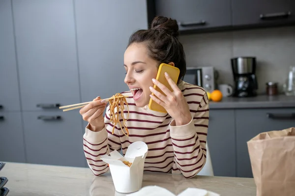 Giovane donna mangiare tagliatelle, parlando sul telefono cellulare — Foto Stock