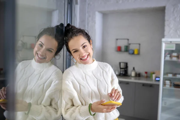 Jonge vrouw die aan de glazen muur staat, mobiel vasthoudend, glimlachend — Stockfoto