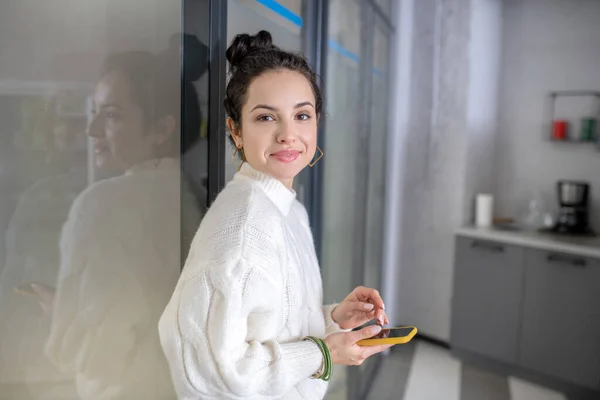 Młoda kobieta opierająca się o szklaną ścianę, trzymająca ruchomy, uśmiechnięty — Zdjęcie stockowe