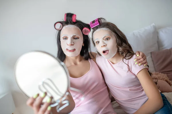 Mamá e hija con mascarilla facial en la cara mirándose en el espejo — Foto de Stock
