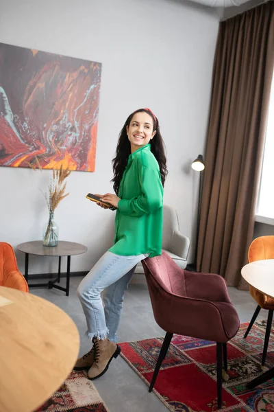 Jovem mulher com smartphone em pé na sala, inclinando-se na cadeira . — Fotografia de Stock