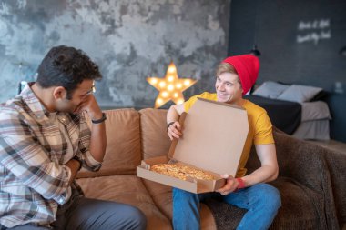 Kırmızı şapkalı genç adam arkadaşına pizza gösteriyor ve gülümsüyor.