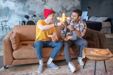 İki genç adam kanepede oturup pizza ve biranın tadını çıkarıyor.