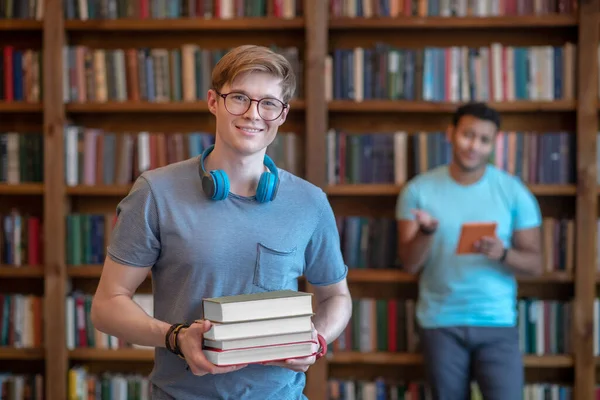 Fair-haired young man in eyeglasses standing in the library with his friend — Stock Photo, Image
