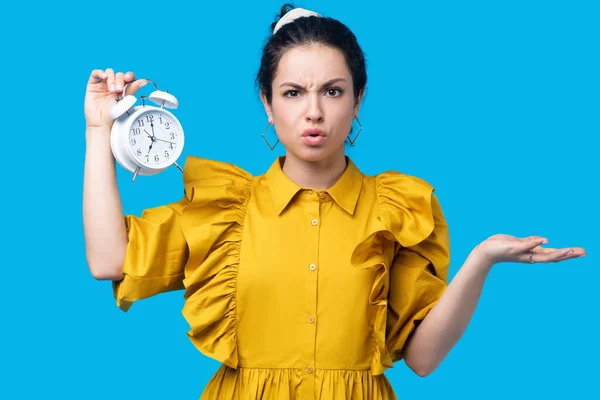 Vrouw in een mosterdjurk met een klok en onzeker kijkend — Stockfoto