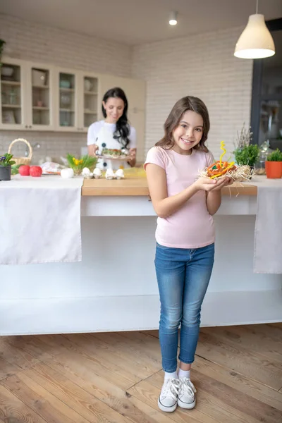 A lány farmerban áll a konyhában és húsvéti zsemlét tart a kezében. — Stock Fotó