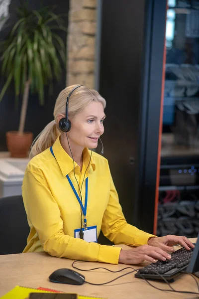 Blonde woman sitting in front of computer in headphones, typing — Stock Photo, Image