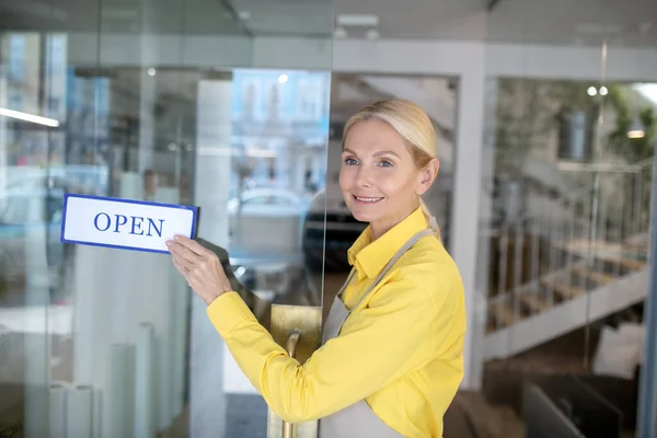 Szőke nő áll a bejáratnál, kezében felirat, hogy nyitott — Stock Fotó