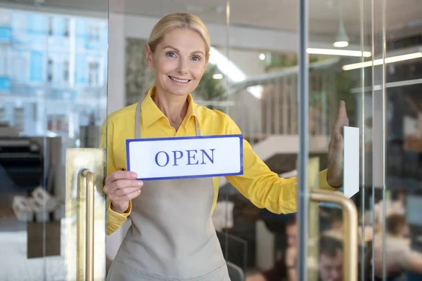 Blondynka otwierająca szklane drzwi, trzymająca znak "otwarte" — Zdjęcie stockowe