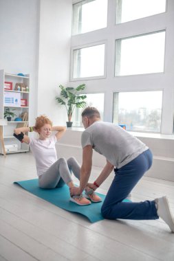 Kadın hasta mekik çekiyor, erkek fizyoterapist ayak bileklerini tutuyor.