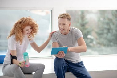 Kadın hasta ve erkek fizyoterapist pencere kenarında oturuyor, tablete bakıyor, tartışıyor, gülümsüyor.