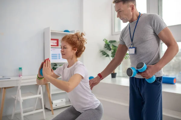 Mannelijke fysiotherapeut houden halters, assisteren vrouwelijke patiënt met hurken — Stockfoto