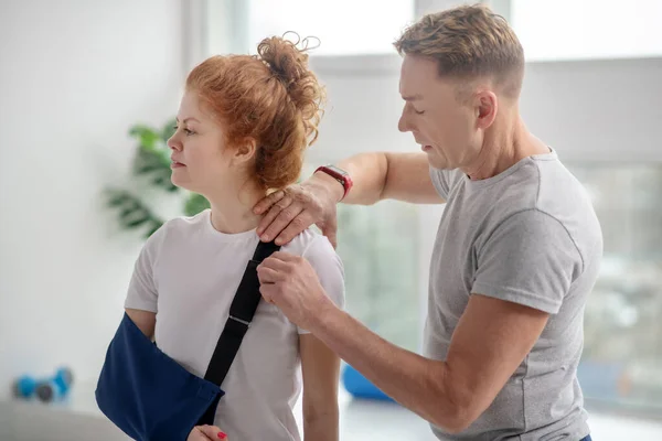 Мужской физиотерапевт осматривает плечо пациентки с повязкой на руке — стоковое фото