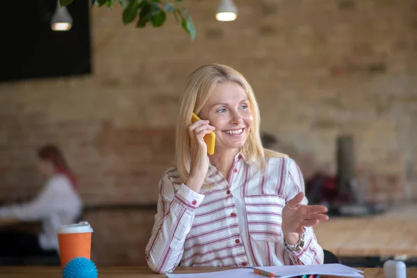 Ξανθιά γυναίκα κάθεται στο γραφείο, έχει τηλεφωνική συνομιλία, χαμογελώντας — Φωτογραφία Αρχείου