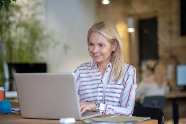 Szőke nő ül az asztalnál, gépel a laptopon, mosolyog — Stock Fotó