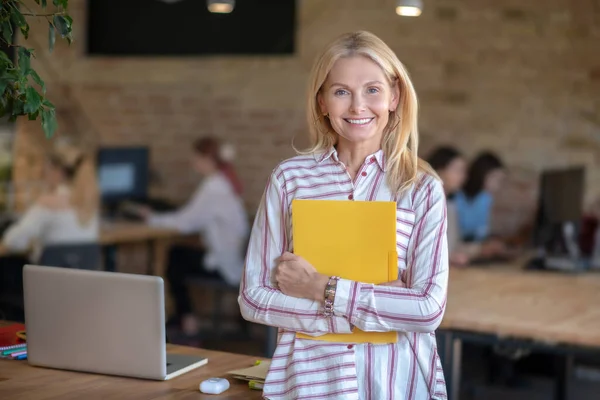 Szőke nő áll az irodában, kezében sárga mappa — Stock Fotó