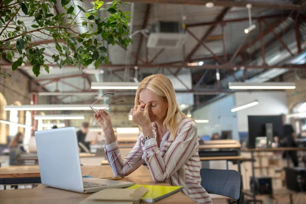 Mujer rubia sentada en el portátil, masajeando su puente nasal, sosteniendo gafas — Foto de Stock