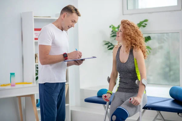 Male physiotherapist making notes, female patient sitting with crutches — Stock Photo, Image