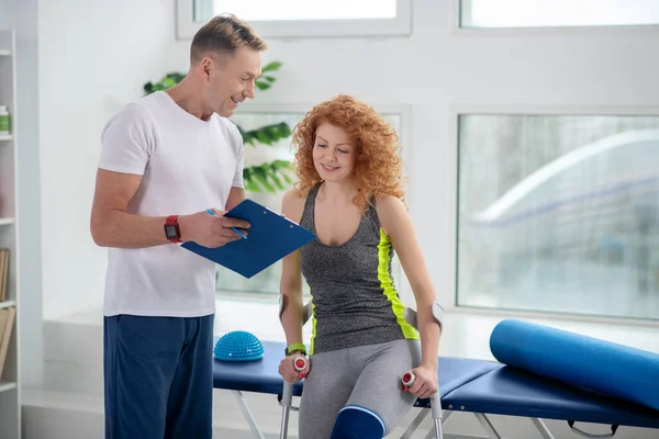 Male physiotherapist showing notes to female patient sitting with crutches — Stock Photo, Image
