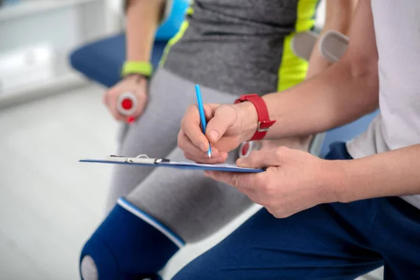 Mężczyzna fizjoterapeuta ręce robi notatki, kobieta pacjent siedzi obok niego — Zdjęcie stockowe