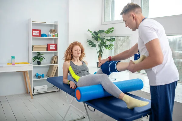 Fisioterapeuta masculino tratando rodilla de paciente mujer acostada en sofá — Foto de Stock