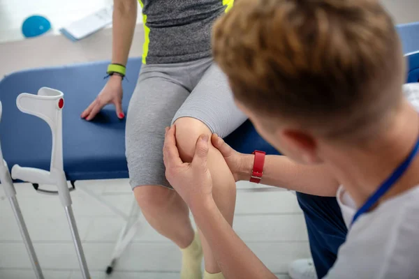 Mužské fyzioterapeutické ruce léčí zranění kolena pacientky sedící na gauči — Stock fotografie