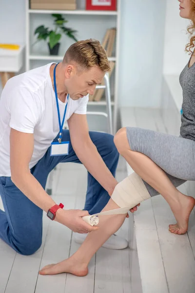 Αρσενικός φυσιοθεραπευτής γονατίζει στο ένα γόνατο, απλώνοντας επίδεσμο στο γυναικείο γόνατο ασθενούς — Φωτογραφία Αρχείου