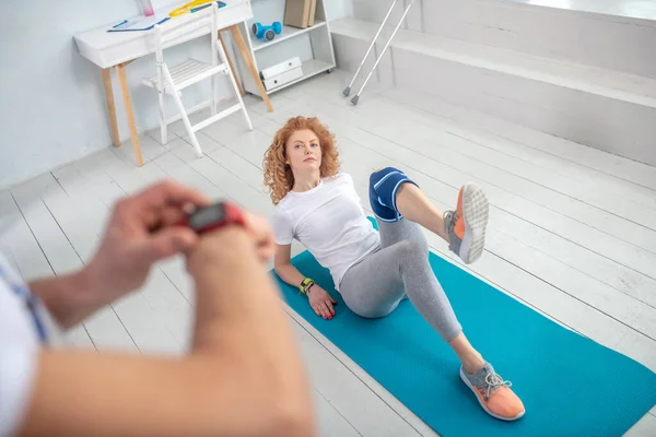 Vrouwelijke patiënt liggend op mat, buigen van haar been, mannelijke fysiotherapeut handen opname tijd met stopwatch — Stockfoto