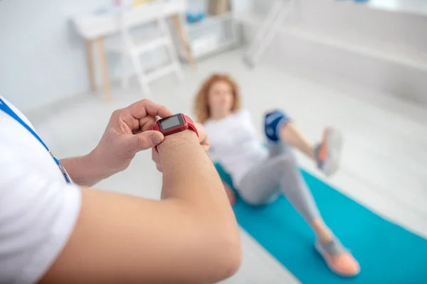 Paciente femenina acostada en la estera, doblando la pierna, manos fisioterapeutas masculinas grabando el tiempo con cronómetro — Foto de Stock