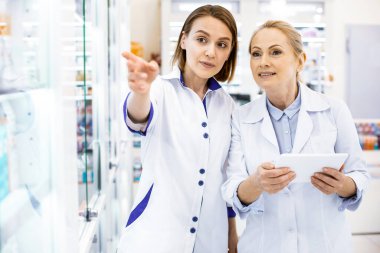 Düşünceli genç kadın eczacılar eczanede vitrini inceliyor.