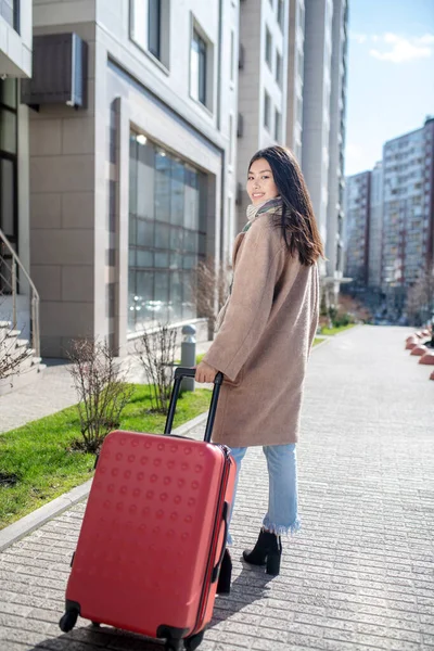 Темноволоса молода жінка в бежевому пальто з червоною валізою — стокове фото