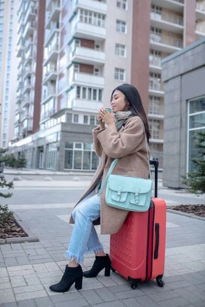 Tmavovlasá mladá žena v béžovém kabátě sedící na svém kufru — Stock fotografie