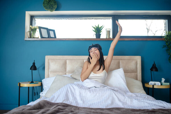 Молодая женщина в маске для сна зевает и растягивается в постели
