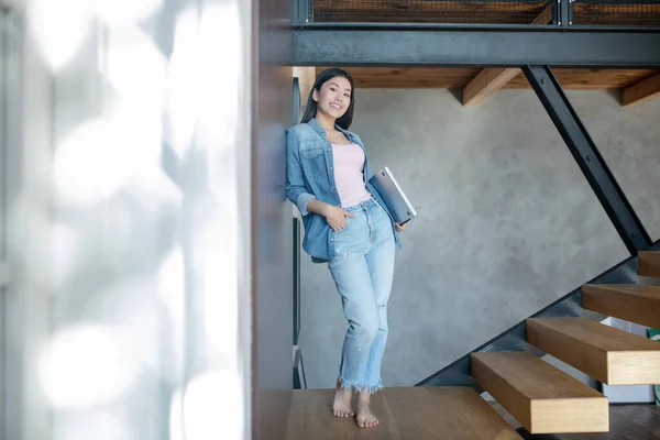 Fiatal nő áll a lépcsőn, tartja laptop a hóna alatt, támaszkodik a falra, mosolyog — Stock Fotó