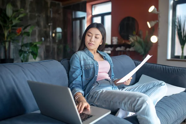 Fiatal nő ül a kanapén, kezében papírok, gépelés a laptopon — Stock Fotó