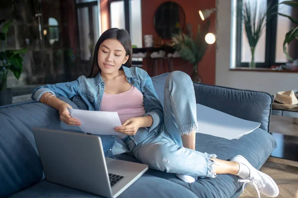 Mladá žena sedí na pohovce s laptopem, drží papíry, čte — Stock fotografie