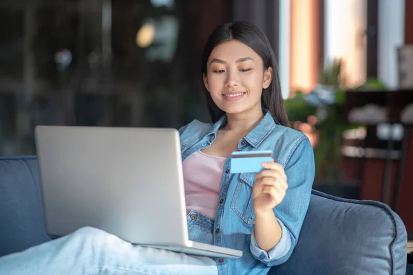 Young woman sitting on sofa with laptop on her knees, holding card