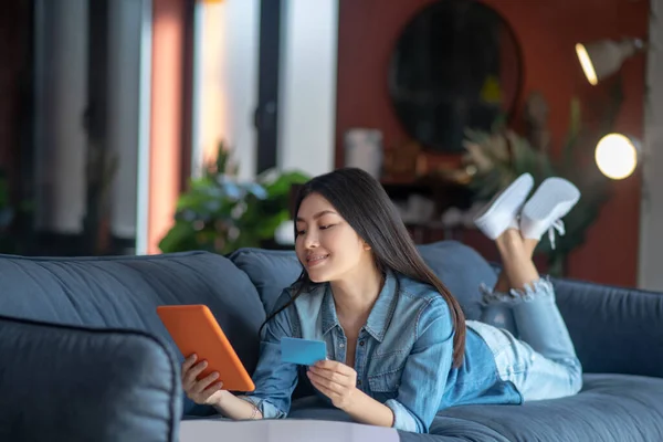 Junge Frau liegt auf Sofa, hält Tablet und Karte in der Hand, blickt auf Bildschirm, die Beine gekreuzt — Stockfoto